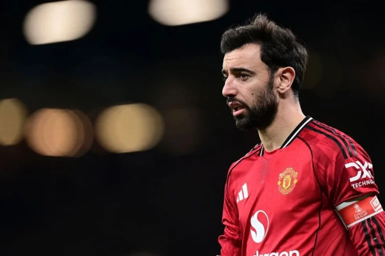 Bruno Fernandes berjanggut dengan seragam merah Manchester United sponsor DXC Technology, berdiri di lapangan malam hari dengan latar belakang lampu sorot